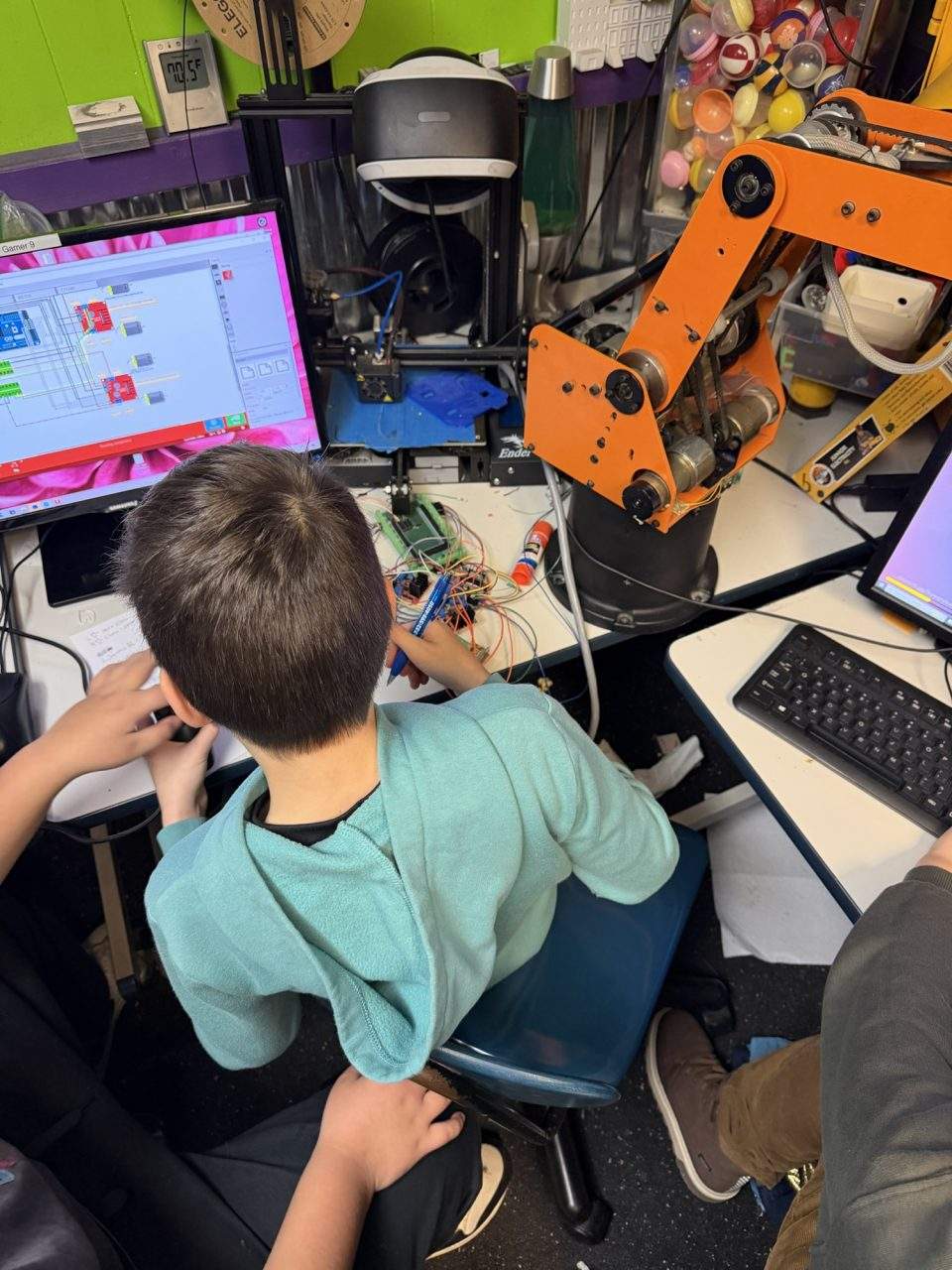 Student programming SCORBOT robotic arm with Scratch on screen next to 3D printer at Plum Crazy Robotics